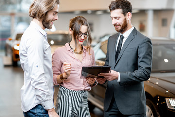 Handsome male dealer selling to a young couple clients a new car, standing together with some documents in the showroom