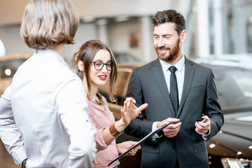 Handsome male dealer selling to a young couple clients a new car, standing together with some documents in the showroom