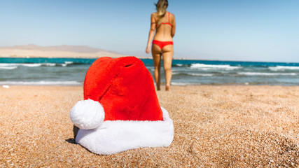 Closeup image of Santa Claus hat lying on the sand at sea. Concept of travel and tourism on Christmas, New Year and winter holidays.