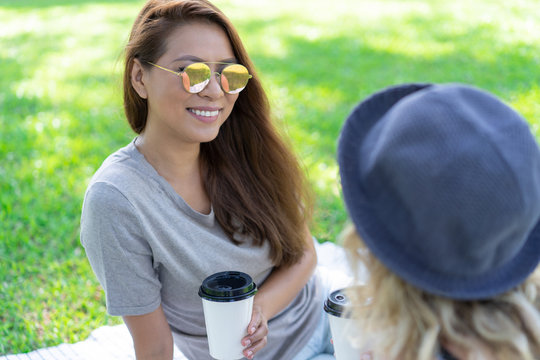 Smiling Asian girl with coffee cup chatting with close friend at picnic. Happy young woman in sunglasses enjoying leisure with friend outdoors. Communication concept