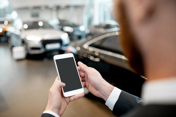 Businessman holding phone with empty screen to copy space in the showroom with cars on the background