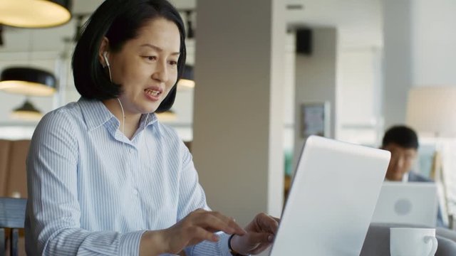 Tracking medium shot of confident Asian woman using laptop computer and earphones for video conversation with colleague