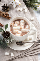 Fototapeta na wymiar Cup of hot chocolate with marshmallow cinnamon sticks on the white wooden table. Winter cocoa drink on a napkin, Christmas-tree golden stars decorations, branches and cones