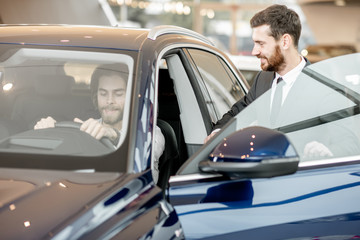 Salesperson helping young man to choose a new car sitting on the driver seat of a luxury car in the showroom