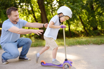 Middle age father showing his toddler son how to ride a scooter in summer park. © Maria Sbytova