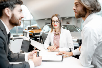 Young business couple signing some documents at the table with salesperson or manager buying or renting car in the showroom