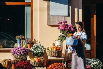 Young female florist seller of flowers and gifts shop waiting for customer on the threshold of her...