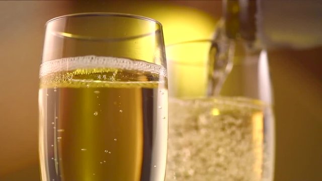 Champagne pouring from a bottle. Two flutes with sparkling wine over golden holiday background. Celebration. Slow motion 4K UHD video footage. 3840X2160