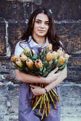 Portrait of charming woman florist in apron with bouquet of protea flowers outdoor over dark background.