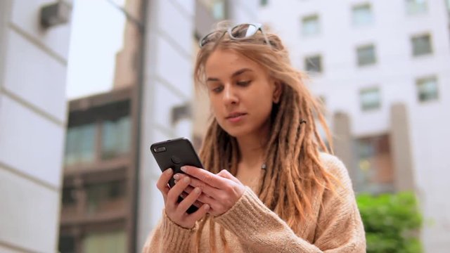 portrait hipster girl with dreadlocks using smartphone on the street downtown area slow motion