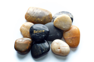 A gray polished river stone with the word believe in gold on a pile of many polished rocks. Over...