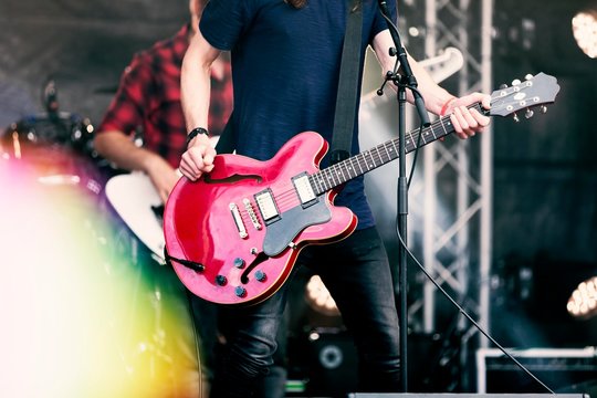 Rock Band Fronted By Heavy Metal Singer In Black T Shirt And Jeans Playing A Red Electric Guitar On Stage At A Festival