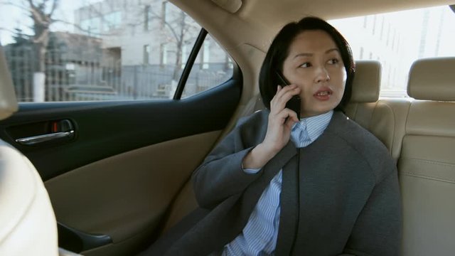 Medium shot of middle-aged Asian female entrepreneur sitting in backseat of car and talking on phone