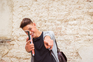 Portrait of the smiling young man who is standing outdoor near the old house wall and holding the notebooks in hand and showing forward by the forefingers