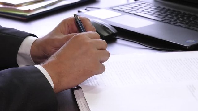 Hand of businessman in suit filling and signing with silver pen partnership agreement form clipped to pad closeup. A businessman is worried and hesitates to sign a document or not.