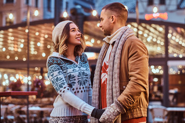 A young romantic couple holding hands and looking at each other while standing on the street at Christmas time, enjoying spending time together.