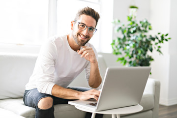 A Young attractive guy is browsing at his laptop, sitting at home on the cozy beige sofa at home, wearing casual outfit