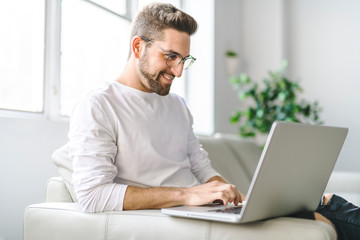 A Young attractive guy is browsing at his laptop, sitting at home on the cozy beige sofa at home, wearing casual outfit