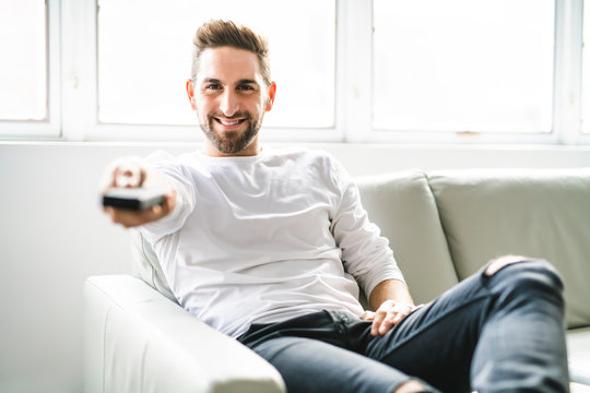 A Happy young man sitting with TV remote control on a couch at home