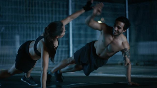 Smiling Happy Athletic Fitness Couple Doing Push Up Exercises and Giving High Five. Workout is Done in a Fenced Outdoor Basketball Court. Night Footage After Rain in a Residential Neighborhood Area.