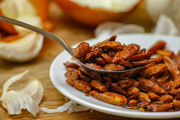 Dried red hot small chilli peppers in spoon and on small ceramic plate. Old wooden table and various of vegetable in the background.