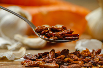 Dried red hot small chilli peppers in spoon and on small ceramic plate. Old wooden table and various of vegetable in the background.