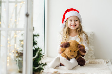 Little girl in the hat of Santa Claus with a plush deer sitting on the window. A child looks out the window and is waiting for Christmas, Santa Claus