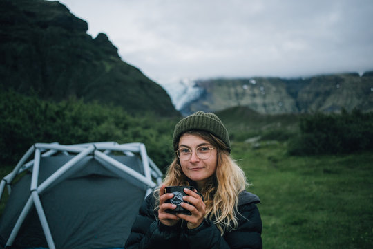 Young beautiful blonde woman or girl looks and smiles at camera at camping site next to tent, epic mountain and glacier on background, holds metal cup or mug with coffee or tea, outdoor lifestyle