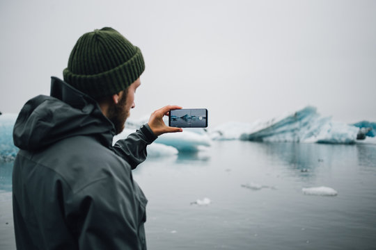 Young man in warm winter clothing stands on beach in iceland or greenland and makes photo on smartphone of icebergs and glaciers floating in water, slowly melting and raising ocean level