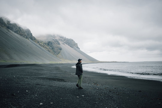 Grey palette image of sandy volcanic beach on cloudy and gloomy day, impressive epic mountains in distance, young man in jacket walks alone towards water, concept lonely or adventure in iceland