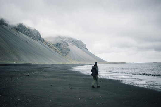 Grey palette image of sandy volcanic beach on cloudy and gloomy day, impressive epic mountains in distance, young man in jacket walks alone towards water, concept lonely or adventure in iceland