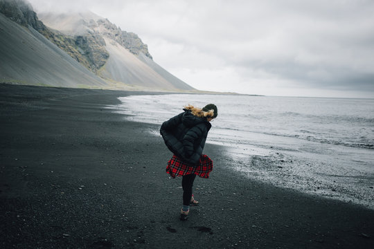 Young woman in cool outdoor outfir, red shirt and puffy down jacket walk in empty cold grey beach with black volcanic sand. Concept explore more and unconventional travels in iceland