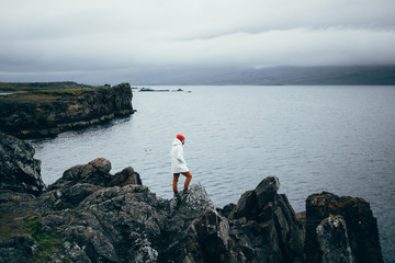 Fisherman or sailor in white raincoat or jacket stands on edge of cliff or rocks overlooks sea or ocean on cloudy rainy day. Moody grey image of icelandic lifestyle . color in greyness - Powered by Adobe