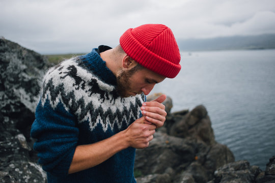 Young handsome man with beard, in trendy blue traditional sweater and red beanie, sailor or fisherman look, lights up cigarette on cloudy day on rocky beach. Hipster or authentic millennial