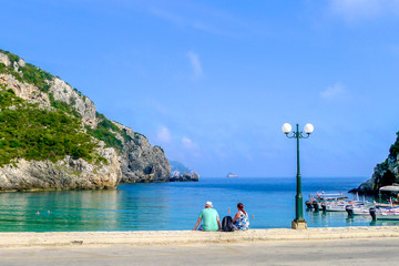 Obraz premium A couple of tourists sit on the waterfront overlooking the turquoise blue waters of Paleokastritsa beach in on the Greek island of Corfu, also known as Kerkyra, in the Ionian sea