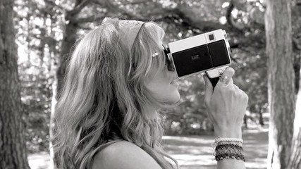 Groovy, young, retro blonde woman films with an 8mm movie camera. Profile shot. She brings camera to eye and then tilts up and down. Black and white.