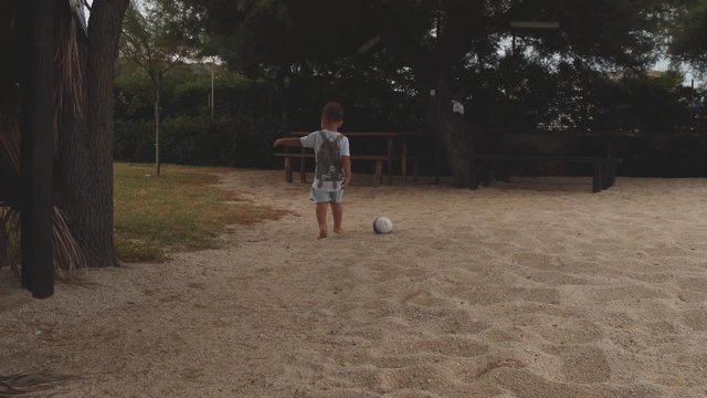 Little boy kicks a ball. Playing the game on sandy beach near park trees. Slow motion