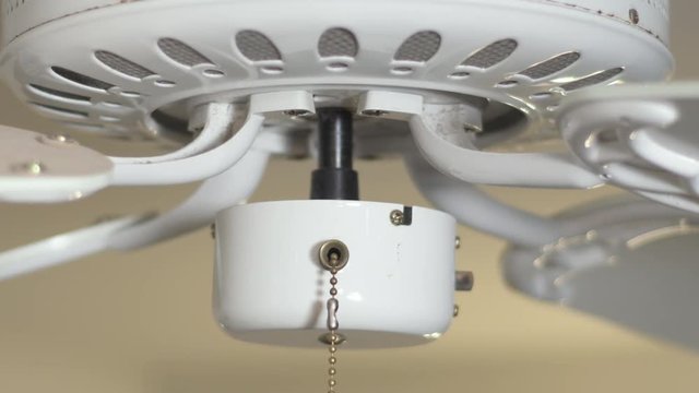 Close up on a white ceiling fan with it being activated via the pull cord, and starts spinning at full speed.