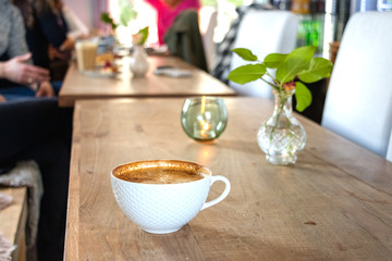 A white cup of cappuccino coffee on a large wooden table with people in the background.