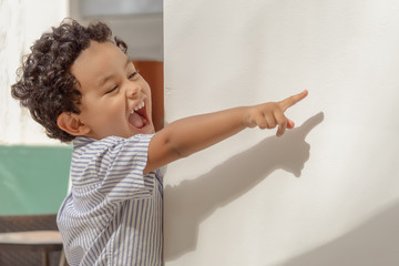 A curly hair little boy looks at a distance behind a white wall with a big laugh pointing his finger.