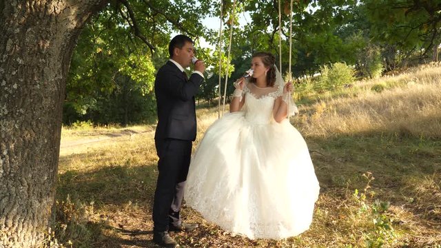 Happy groom in suit and bride in white dress drink champagne from beautiful wine glasses and ride on swing in park smiling to each other. Couple in love drink wine.