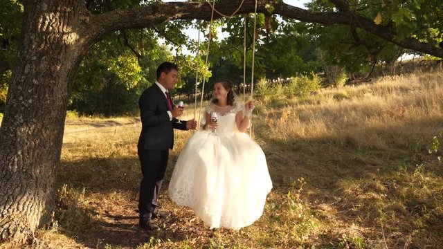 Happy groom in suit rolls on swing bride in white dress drink champagne from beautiful wine glasses in park smiling to each other. Couple in love drink wine.