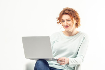 Fototapeta premium Attractive red haired mature smiling woman sitting in the chair with a laptop on her knees, working or watching something against white background