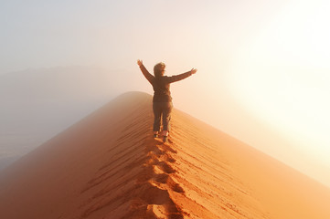 Person standing on top of dune in desert and looking at rising sun in mist with hands up, travel in...