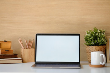 Close-up mockup laptop on white table. books, coffee, pencil. In the background a wood wall and house plants. Empty workplace.