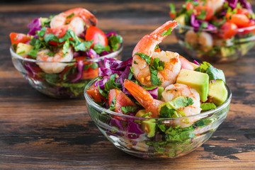 Mexican salad with fried shrimps, fresh avocado, cherry tomatoes, coriander and red cabbage in glass bowl on wooden rustic table, close-up