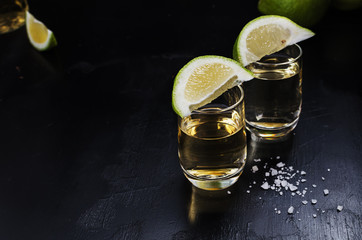 Golden tequila shots with sea salt, juicy lime on vintage black wooden board, toned, selective...
