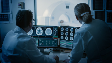 In Control Room Doctor and Radiologist Discuss Diagnosis while Watching Procedure and Monitors Showing Brain Scans Results, In the Background Patient Undergoes MRI or CT Scan Procedure. - obrazy, fototapety, plakaty