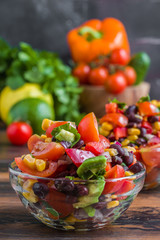 Vegetarian salad in Mexican style, made from avocado, beans, corn, red onions, tomatoes and cilantro, in glass bowls on a wooden table.