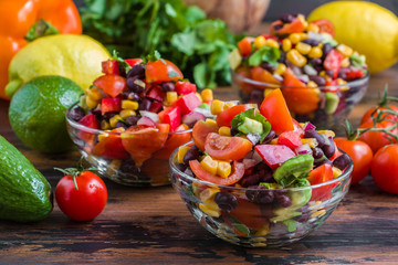 Mexican salad with avocado, tomatoes, beans, corn, red onions and cilantro in glass bowls on a wooden table.